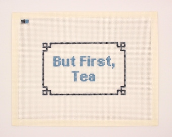Op de afbeelding: Een crème-kleurig borduurcanvas met de tekst "But First, Tea" in lichtblauwe letters, omlijst door een zwarte rand. Het canvas heeft een rastertextuur, wat duidt op een handgemaakt item.