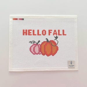 Könnte beinhalten: Eine weiße Stickleinwand mit dem orangefarbenen Text HELLO FALL über zwei Kürbissen. Ein Kürbis ist rosa und der andere orange, beide mit schwarzen Stielen und Ranken. Ein kleiner Farbpalettenstreifen befindet sich oben links und ein Blue Willow Logo-Etikett unten rechts.