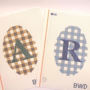 Può includere: Due tele per punto croce con disegni a forma di uovo. Uno presenta un motivo a quadri marrone e beige con una "A" verde, l'altro ha un motivo a quadri blu e bianco con una "R" blu. Le lettere e i quadri sono cuciti su sfondo bianco.
