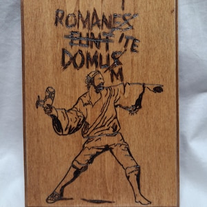 Puede incluir: Placa de madera con diseño pirograbado. La obra representa una figura lanzando un objeto, con el texto "Romanes Eunt Domus" encima. La placa es rectangular y está hecha de madera clara.