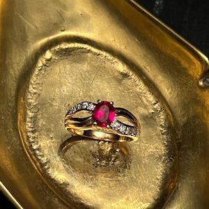 May include: A gold ring with a large oval ruby center stone and smaller clear stones. The ring is set in a split-shank design and sits on a gold-colored tray.