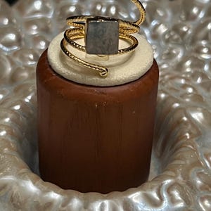 May include: A gold-toned spiral ring featuring a textured band and a central square-shaped gray stone. The ring wraps around the finger area with small spherical accents at each end of the wire, showcasing a metallic finish and a flat stone surface.
