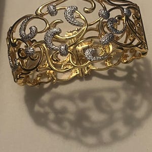 May include: A gold-toned bracelet with an openwork design. The bracelet features a series of swirling, floral-inspired patterns, with small, sparkling accents. The bracelet is a hinged cuff style.