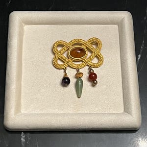May include: A gold-toned brooch featuring a central oval amber-colored stone surrounded by a braided metal frame. Three dangling charms hang from the bottom: a dark purple bead, a pale green tapered bead, and a round red bead with gold accents.