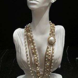 May include: A multi-strand necklace featuring gold-tone chains interspersed with white faux pearls and small gold-tone decorative beads. The design includes a prominent circular gold-tone pendant with a large central white faux pearl.