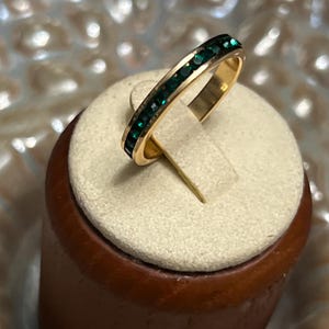 May include: A gold-toned band ring featuring a channel setting of small, square-cut dark green stones that span the top half of the circular metal band. The ring is displayed upright on a circular beige fabric-covered stand with a wooden base.
