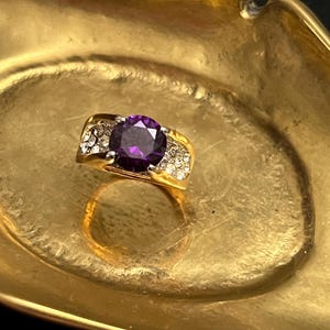 May include: A gold-toned ring with a large, round, deep purple gemstone at the center. The ring is further embellished with smaller, clear gemstones on either side of the central stone. The ring is set in a gold-colored metal and is displayed in a gold-colored dish.
