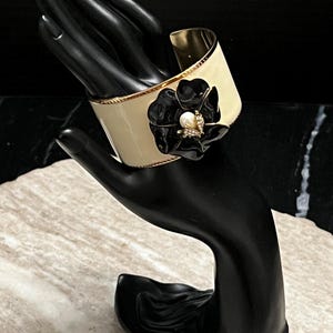 May include: A cream-colored cuff bracelet with a gold interior and trim. A black flower with a pearl and rhinestone center adorns the front. The bracelet is displayed on a black hand-shaped jewelry holder.