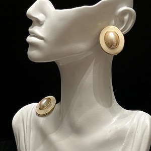 May include: A pair of round earrings featuring a central oval faux pearl surrounded by a smooth cream-colored border and a thin gold-tone metal rim. One earring is displayed on the ear of a white mannequin, while the other rests on the shoulder.