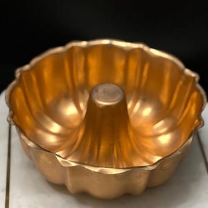 May include: A copper-colored bundt cake pan with a fluted edge and a center tube. The pan is made of metal and has a shiny finish.
