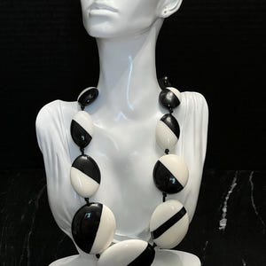 May include: A bold black and white statement necklace. The necklace features large, oval-shaped beads in alternating black and white sections. The necklace is displayed on a white mannequin bust against a black background, highlighting the jewelry's design.