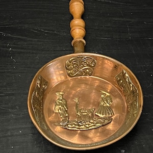 May include: A copper-colored pan with a wooden handle. The pan features embossed figures of people and an animal, with decorative floral accents. The handle is made of turned wood. A vintage kitchenware item.
