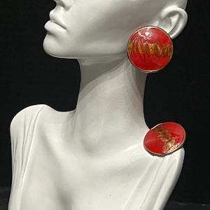 May include: Red and gold round earrings with a gold trim. The earrings have a textured design in gold. The earrings are displayed on a white mannequin head and shoulder form.