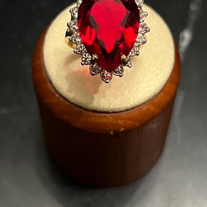 May include: A gold ring featuring a large, pear-shaped, deep red gemstone as the centerpiece. The central stone is surrounded by a halo of small, clear, round-cut stones set in a scalloped metal frame, creating a bright, reflective appearance.