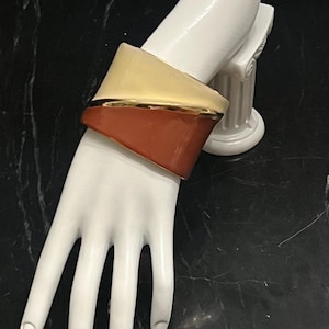 May include: A wide cuff bracelet featuring a two-tone design with a smooth cream-colored upper section and a terracotta-colored lower section, separated by a thin, polished gold-tone metal band that runs diagonally across the center of the piece.