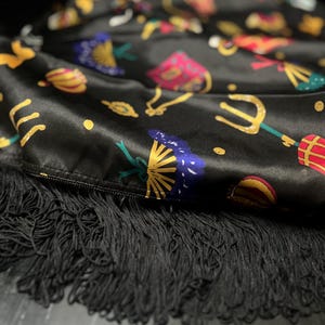 May include: Black satin fabric with a colorful pattern of various shapes and designs. The fabric has a black fringe trim.