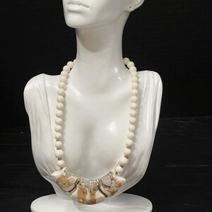May include: A cream-colored beaded necklace with a pendant featuring a mosaic design of beige, brown, and gold tones. The necklace is displayed on a white bust against a black background.