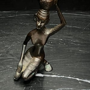 May include: A dark bronze-toned metal figurine depicting a kneeling figure with a smooth, polished finish. The figure is shown in a seated position with one arm raised, balancing a small, textured vessel on top of its head. The sculpture features defined facial features and a simple, stylized form with visible metallic highlights.