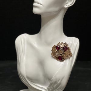 Puede incluir: Busto blanco con una broche dorada. La broche presenta tres piedras preciosas rojas y piedras claras más pequeñas. La broche está engastada en un metal dorado texturizado. El busto es una representación estilizada de la parte superior del cuerpo de una persona.