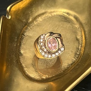 May include: A gold ring with a pink oval gemstone at the center, surrounded by a halo of small, clear gemstones. The ring is set in a gold band with a decorative swirl design. The ring is displayed in a gold-colored dish.