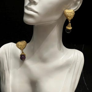 May include: A pair of gold-toned earrings featuring a heart-shaped stud with an intricate embossed scroll pattern. Each earring has a dangling component consisting of a gold-toned conical cap and a smooth, round, deep purple bead at the base.