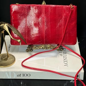 May include: A vibrant red rectangular handbag with a textured, snake-like pattern. The bag has a long, thin red strap and gold-colored hardware. The bag is resting on a book with the text "AD at 100".