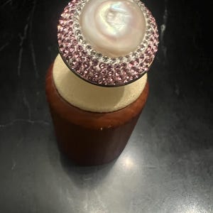 May include: A circular ring featuring a large, iridescent white central pearl surrounded by a border of small, sparkling pink crystals set in a metallic frame. The ring is displayed on a cylindrical, light-colored wooden stand.