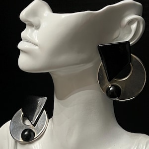 May include: Geometric silver-tone earrings with black accents. Each earring features a circular design with a black triangle and a black sphere. The earrings are displayed on a white bust against a black background.