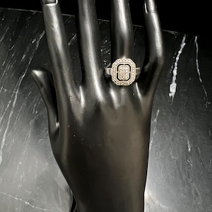 May include: Silver ring with a square, diamond-studded center and a halo of smaller diamonds. The ring is displayed on a black hand-shaped jewelry display, showcasing its intricate design and sparkle.