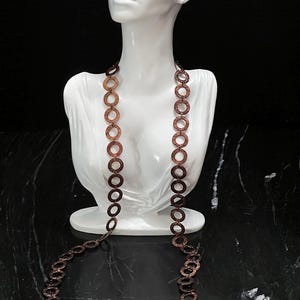 May include: A long necklace composed of numerous interconnected circular links with a textured, metallic copper finish. The chain features a repeating pattern of small, flat, round discs linked together to form a continuous, elongated loop design.