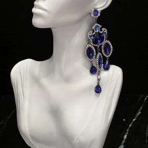 May include: A long, ornate chandelier earring featuring a dark metal setting adorned with multiple deep blue, teardrop-shaped crystals and smaller, clear, round rhinestones arranged in a tiered, intricate, and dangling geometric design.