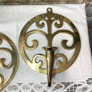 May include: Two brass wall sconces. Each sconce features a circular design with an ornate, openwork pattern and a central, conical candle holder. The sconces have a loop at the top for hanging and are made of shiny brass.
