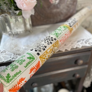 May include: Rolled paper with colorful floral and leaf patterns. The patterns include green, orange, black, and gold designs on a white background. The paper is wrapped in clear plastic, and is likely wallpaper or wrapping paper.