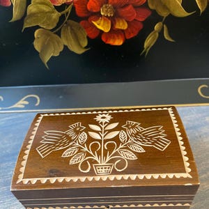 May include: A small, rectangular wooden box with a hinged lid. The lid features a carved design of two birds flanking a floral arrangement. The box is stained a dark brown with white accents and a scalloped edge.