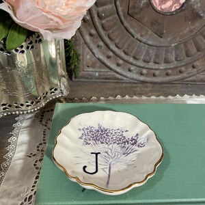 Puede incluir: Un plato peque&ntilde;o, blanco, con borde festoneado y borde dorado. El plato presenta un dise&ntilde;o floral p&uacute;rpura y la letra "J". El plato descansa sobre un libro verde vintage.