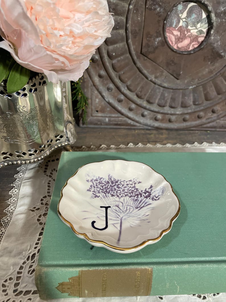 Puede incluir: Un plato peque&ntilde;o, blanco, con borde festoneado y borde dorado. El plato presenta un dise&ntilde;o floral p&uacute;rpura y la letra "J". El plato descansa sobre un libro verde vintage.