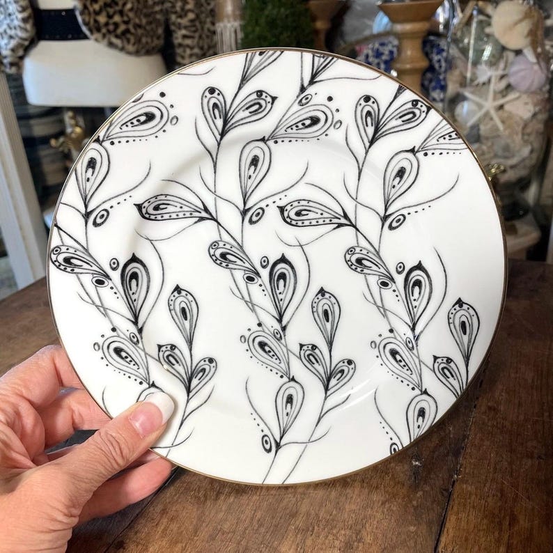 May include: White ceramic plate with a black and gold design. The plate features a black floral pattern with a gold rim. The design is intricate and detailed, with a delicate, hand-drawn aesthetic. The plate is held by a hand.