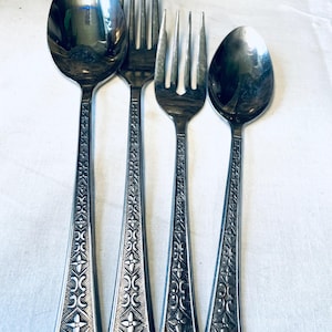 Puede incluir: Un juego de cuatro cubiertos de acero inoxidable: dos tenedores y dos cucharas. La cubertería tiene un diseño floral decorativo en los mangos. Las cucharas y los tenedores están dispuestos en fila sobre una superficie blanca.