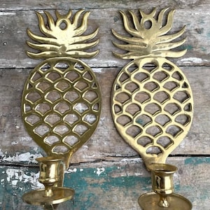 May include: Two brass pineapple-shaped wall sconces. Each sconce features a detailed pineapple design with a candle holder at the base. The sconces are made of gold-colored metal and are mounted on a weathered wooden surface.
