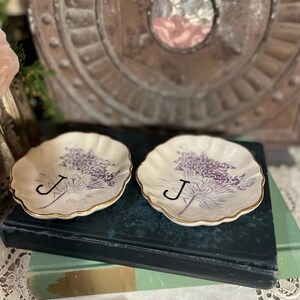 Puede incluir: Dos platos peque&ntilde;os, de color blanquecino, con bordes festoneados y ribete dorado. Cada plato presenta un dise&ntilde;o floral morado y la letra "J". Los platos descansan sobre un libro azul oscuro.