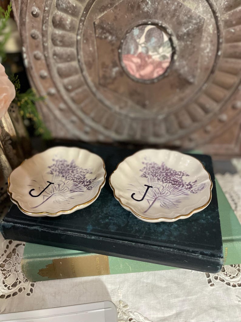 Puede incluir: Dos platos peque&ntilde;os, de color blanquecino, con bordes festoneados y ribete dorado. Cada plato presenta un dise&ntilde;o floral morado y la letra "J". Los platos descansan sobre un libro azul oscuro.