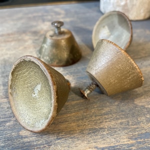 Puede incluir: Cuatro tiradores de armario antiguos de latón con forma cónica y un tornillo pequeño. Los tiradores tienen un aspecto deslustrado y envejecido, lo que sugiere que son vintage o reciclados. Los tiradores están dispuestos sobre una superficie azul-gris desgastada.