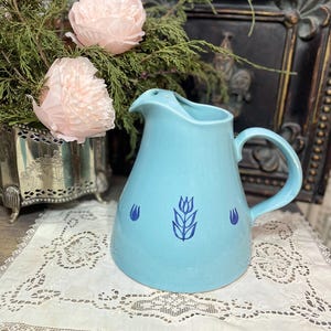 Può includere: Una brocca in ceramica azzurra con manico e beccuccio. La brocca è decorata con un motivo floreale blu scuro. La brocca è appoggiata su una tovaglietta di pizzo bianco. Fiori di carta rosa e verde in un contenitore d'argento sullo sfondo.