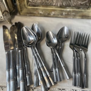 May include: A selection of stainless steel cutlery featuring bamboo-style handles. The set includes knives, spoons, and forks, arranged on a white lace tablecloth. The silverware has a shiny, reflective surface.