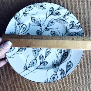 May include: White ceramic plate with a black and white floral design. The plate has a gold rim and is approximately 8 inches in diameter. A wooden ruler is placed across the plate.