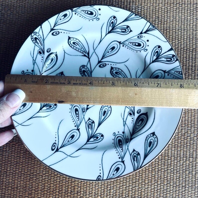 May include: White ceramic plate with a black and white floral design. The plate has a gold rim and is approximately 8 inches in diameter. A wooden ruler is placed across the plate.