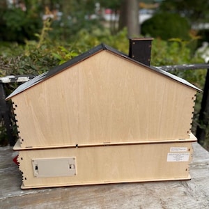 May include: A two-story wooden house-shaped box with a black roof and chimney. The box is made of light-colored wood with visible joinery. A battery compartment is visible on the side. The house is likely a decorative item or a storage box.