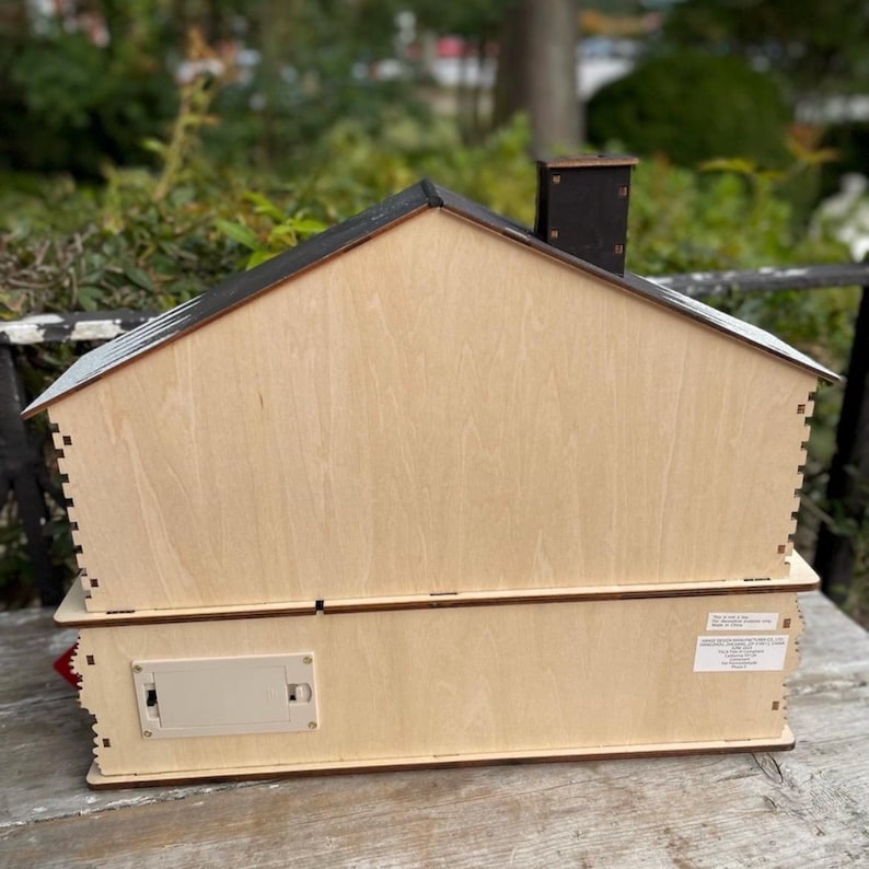 May include: A two-story wooden house-shaped box with a black roof and chimney. The box is made of light-colored wood with visible joinery. A battery compartment is visible on the side. The house is likely a decorative item or a storage box.