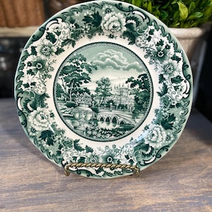 Puede incluir: Un plato decorativo con un diseño floral verde y blanco. El centro presenta un paisaje con un puente y un edificio. El plato se exhibe sobre un soporte de metal dorado. El borde del plato tiene un patrón floral detallado.