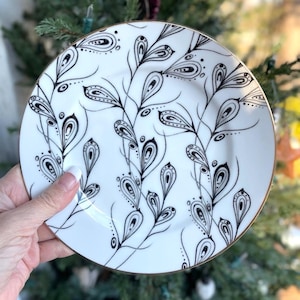 May include: White ceramic plate with a black and gold design. The plate features a repeating pattern of stylized black leaves and a gold rim. The design is hand-drawn and elegant, suitable for serving food or as a decorative piece.